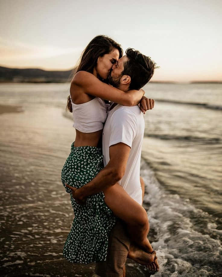 صورة زوجين على الشاطئ يتقبلان بينما يحمل الرجل المرأة وسط الأمواج خلفية
